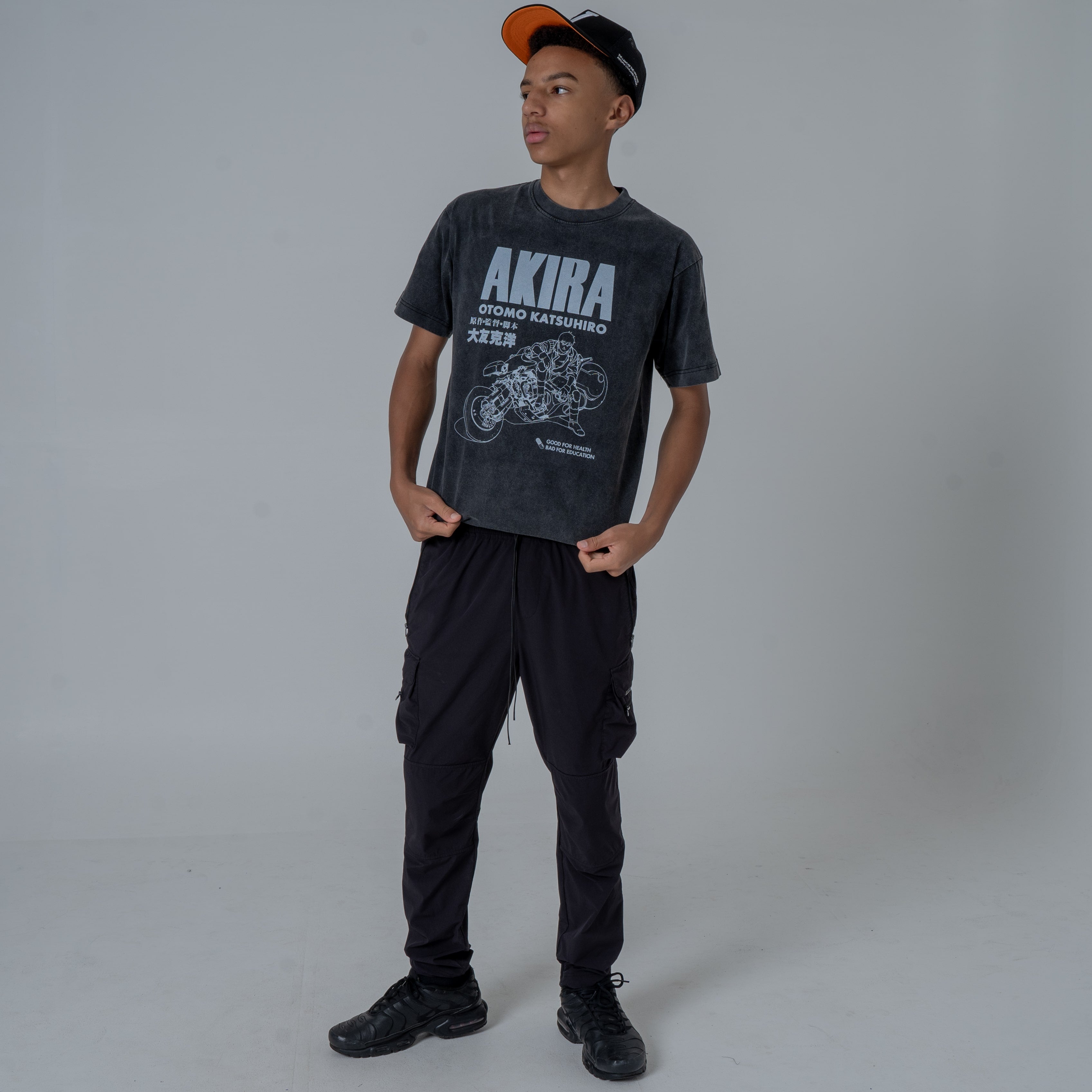 Full body studio shot of a male model looking to the left, wearing an Akira anime T-shirt with a white graphic of Kaneda on his motorcycle, styled with black joggers and black sneakers.