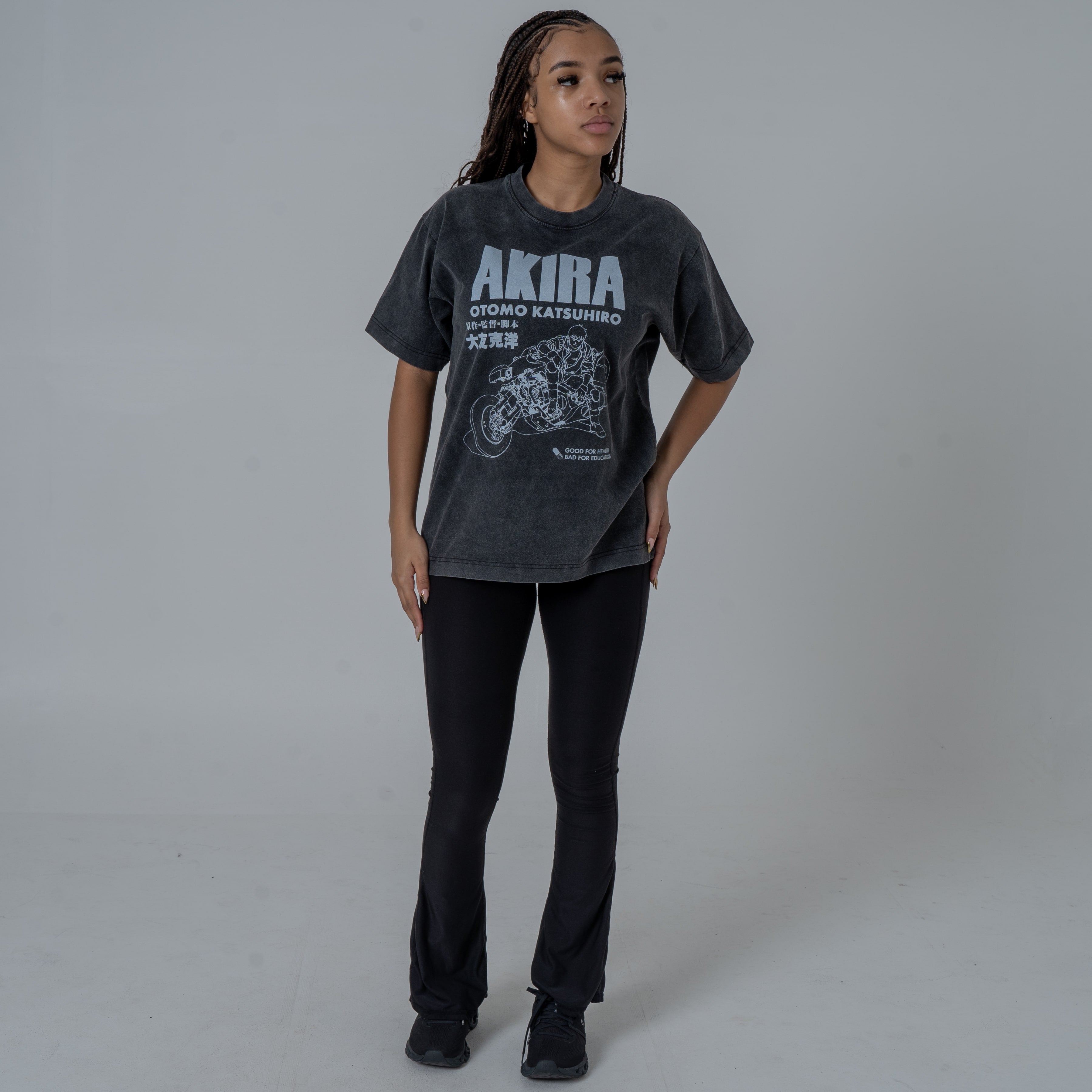 Full body shot of a female model in a studio wearing a black vintage-washed anime T-shirt featuring a white Akira graphic with Kaneda riding his iconic motorcycle, paired with black flared pants and black sneakers