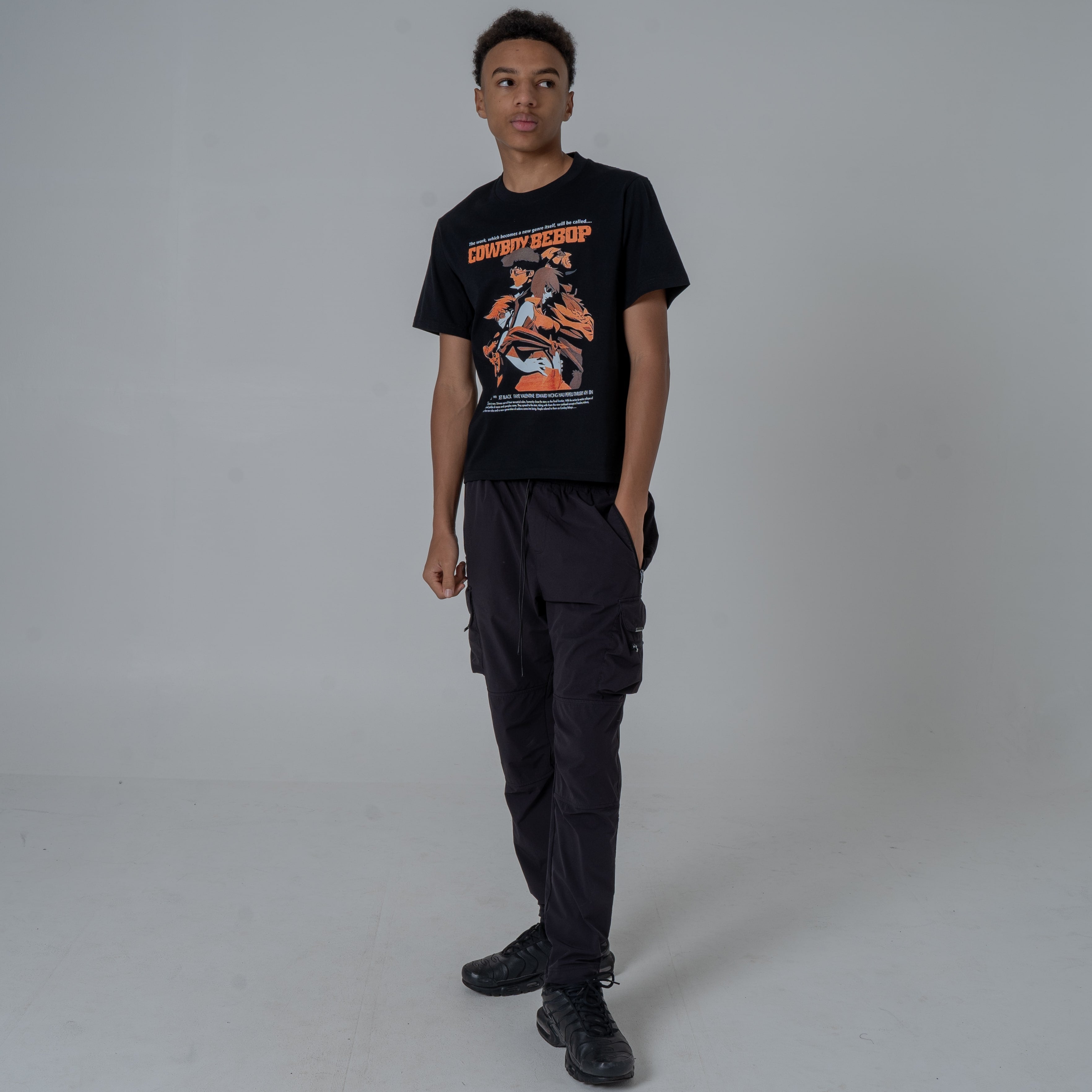 Full body shot of a male model in a studio wearing a black anime T-shirt with an orange Cowboy Bebop graphic featuring Ein, Spyke, Jet, Ed and Faye while looking right.