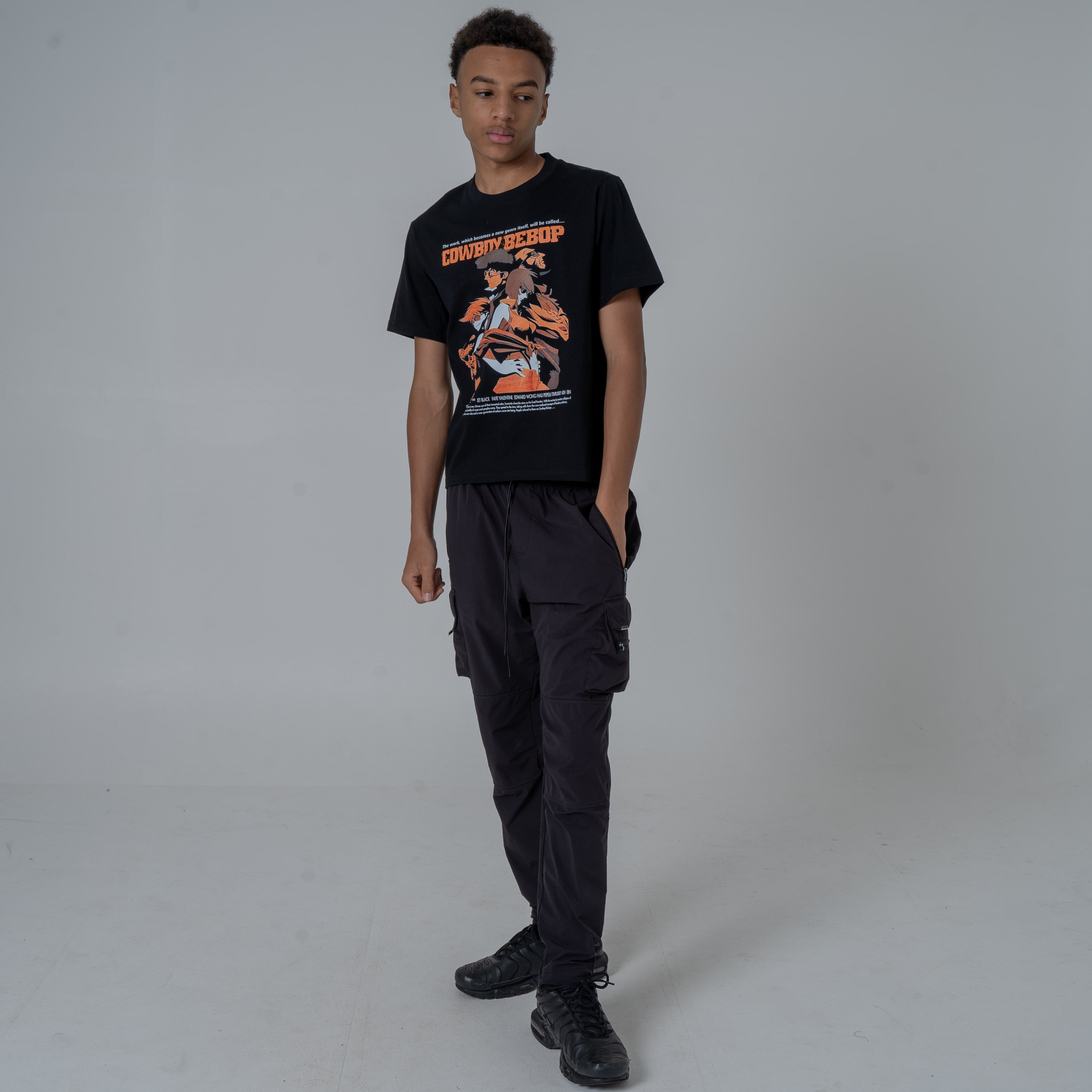 Full body shot of a male model in a studio wearing a black anime T-shirt with an orange Cowboy Bebop graphic featuring a the main characters including spyke paired with boots and joggers.