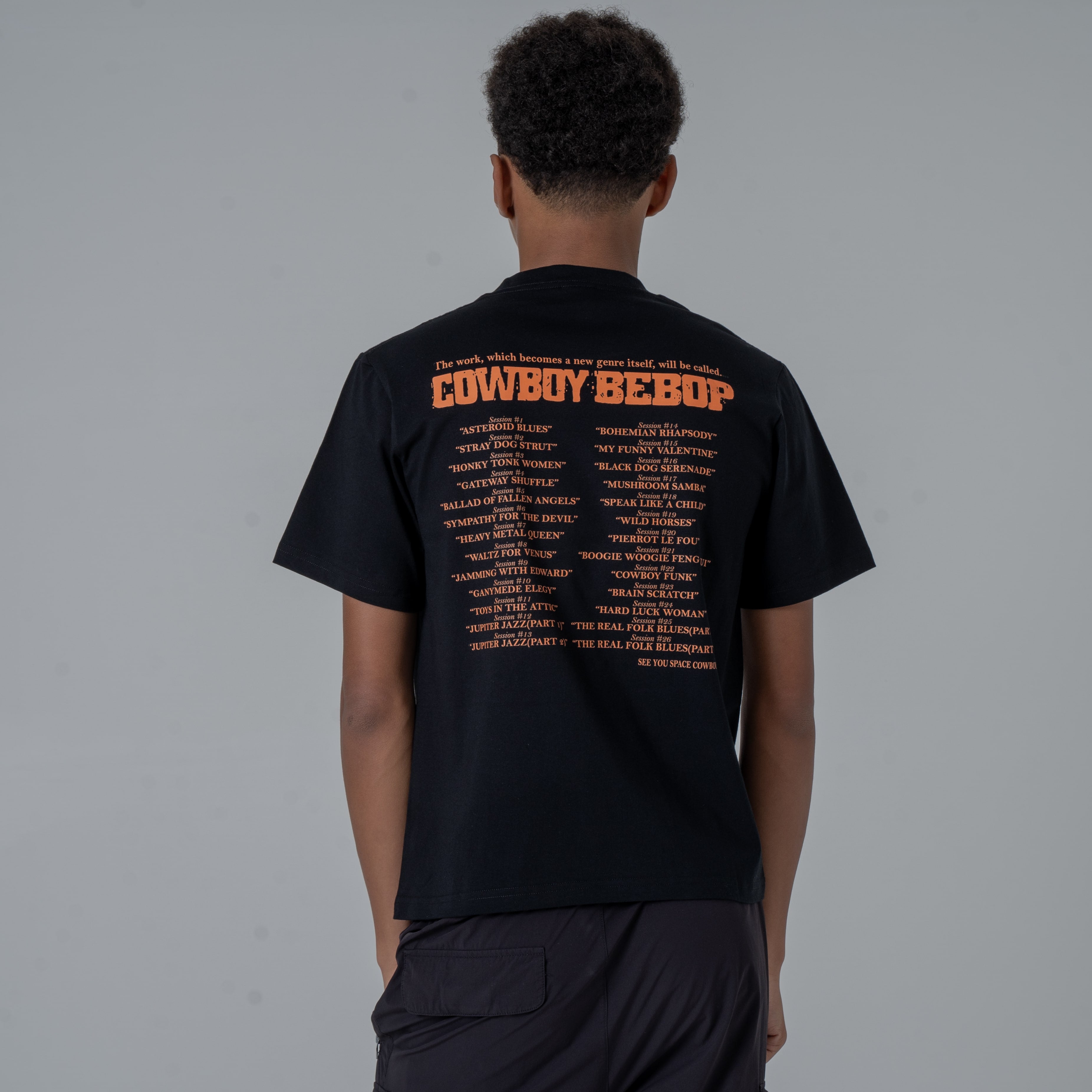 Back view of a male model in a studio wearing a black anime T-shirt with an orange Cowboy Bebop graphic featuring a list of episode titles in vintage-style.
