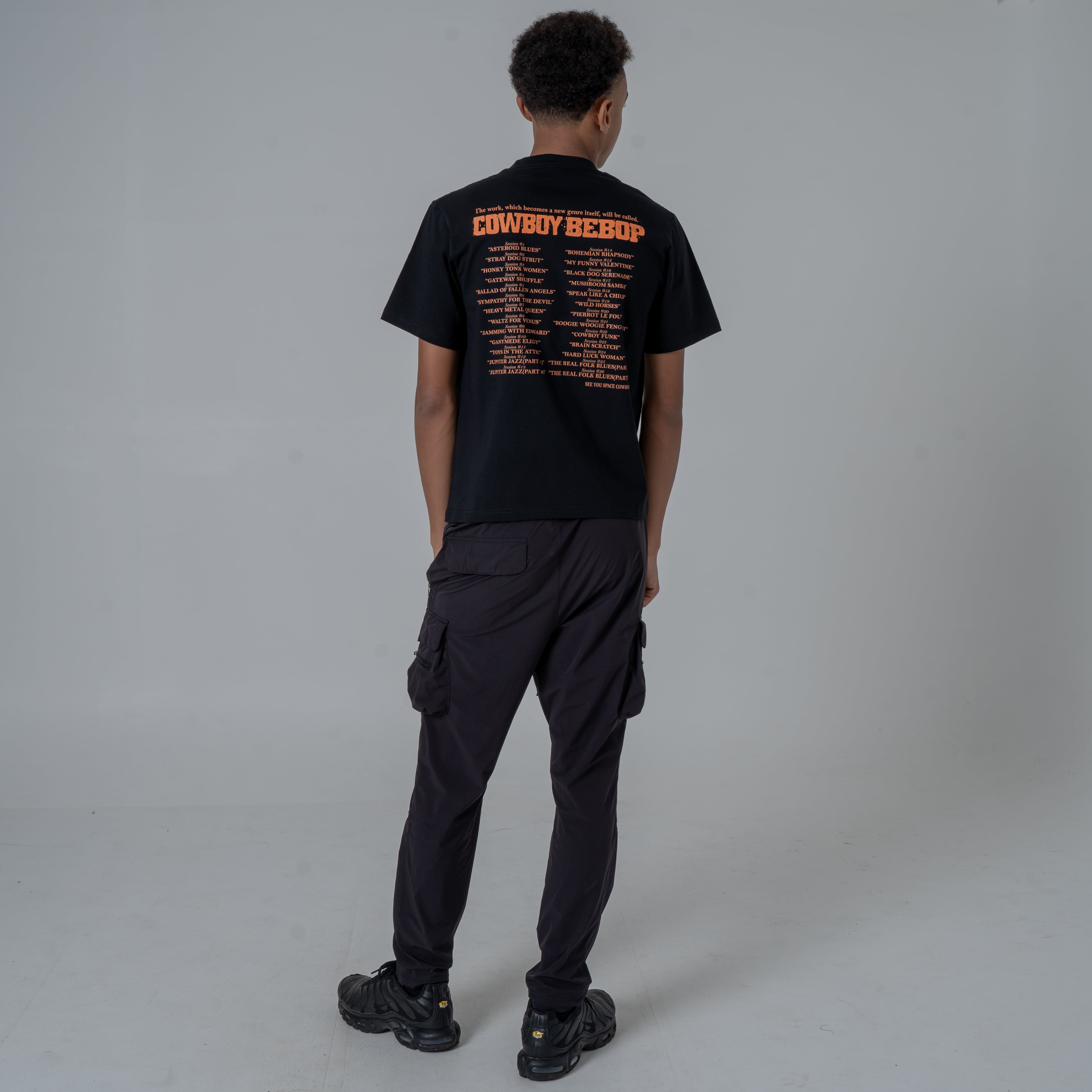 Full body shot of the back view of a male model in a studio wearing a black anime T-shirt with an orange Cowboy Bebop graphic featuring a list of episode titles in vintage-style paired with joggers and sneakers.