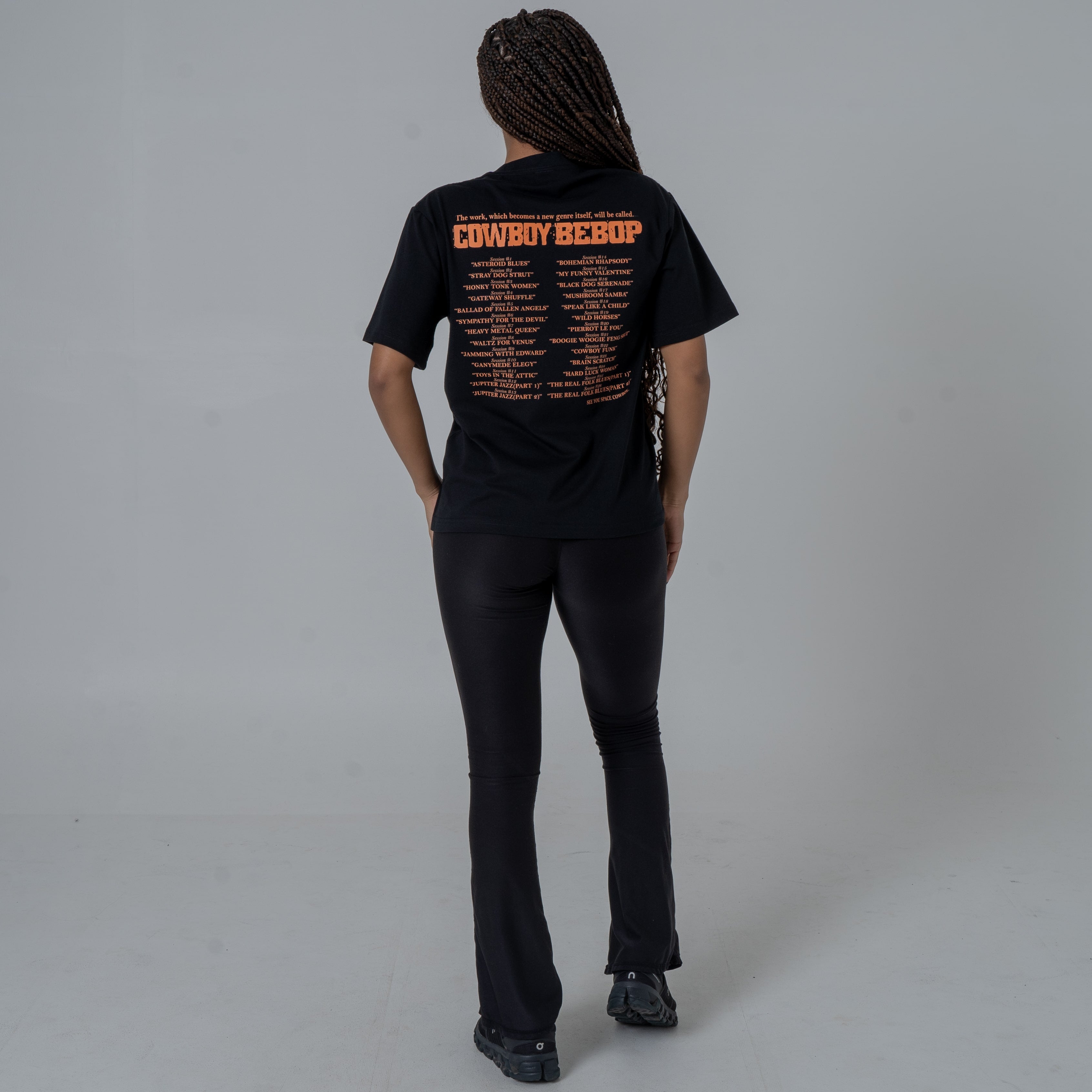 Full body shot of the back view of a female model in a studio wearing a black anime T-shirt with an orange Cowboy Bebop graphic featuring a list of episode titles in vintage-style .