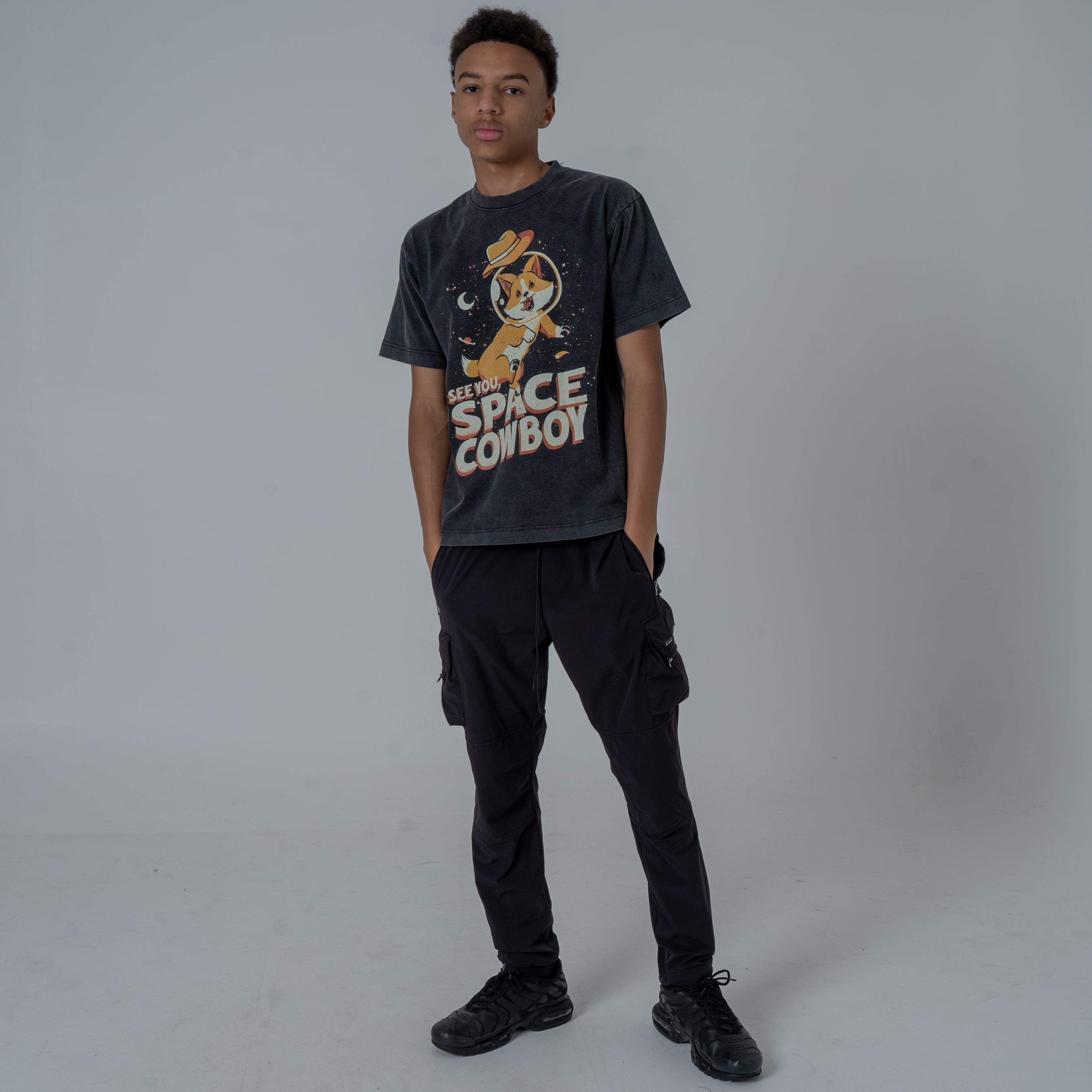 Male model with both of his hand tucked in his pants wearing a vintage-style black T-shirt featuring Ein, the dog from Cowboy Bebop, in a space helmet with the text 'See You Space Cowboy,' styled with black flared pants and black sneakers.