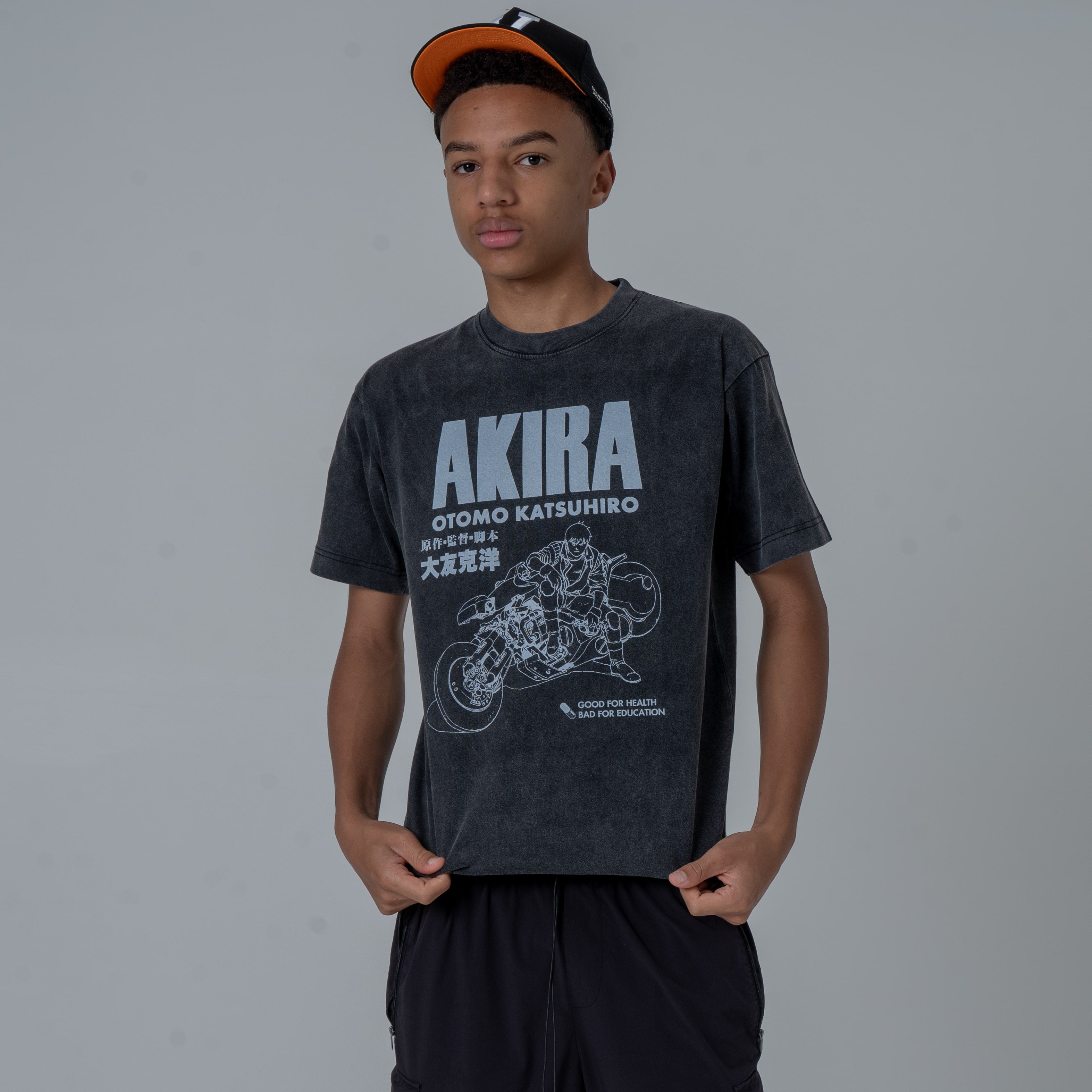Male model in a studio wearing a black vintage-washed anime T-shirt featuring a white Akira graphic with Kaneda riding his iconic motorcycle, paired black and orange colored cap.