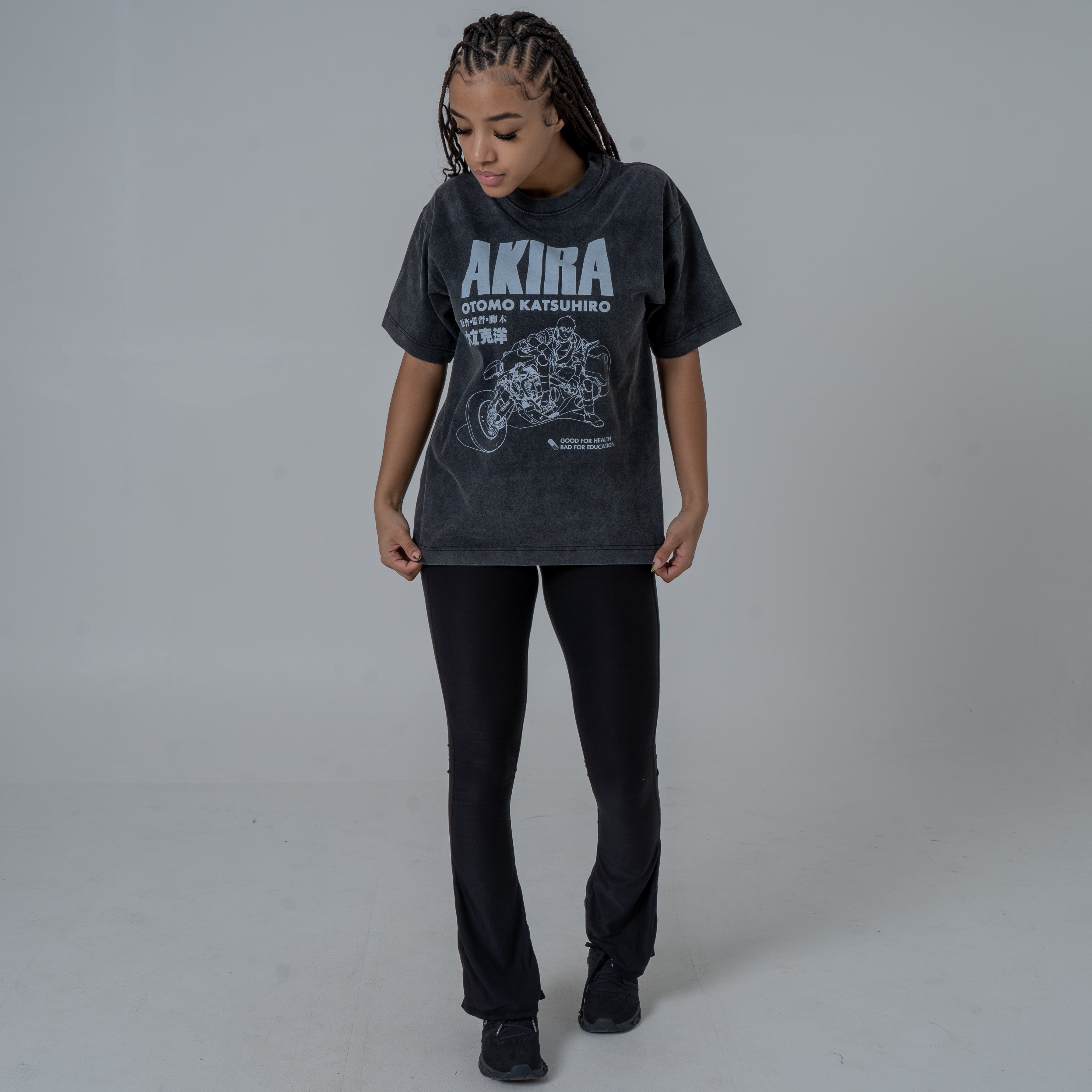 Female model in a studio wearing a black vintage-washed anime T-shirt featuring a white Akira graphic with Kaneda riding his iconic motorcycle, paired with black flared pants and black sneakers